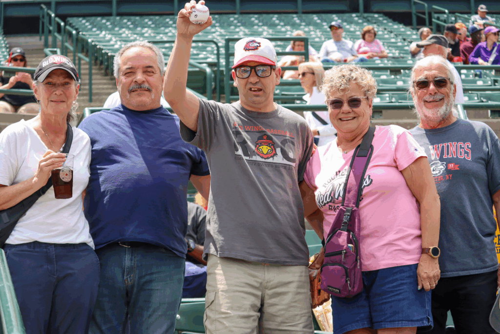 Individuals smiling and standing together at baseball game, with one individual in the center holding up a baseball